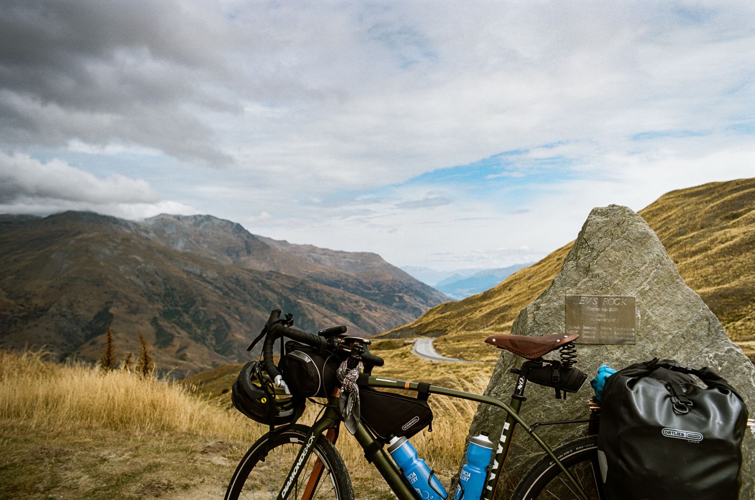 Loaded touring bike leaning against a rock overlooking a panoramic view of the New Zealand countryside with a road winding down the mountain in the distance.