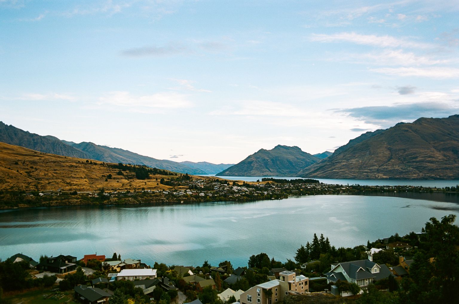 View of Lake Wakatipu surrounded by houses and evergreens with mountains in the distance.