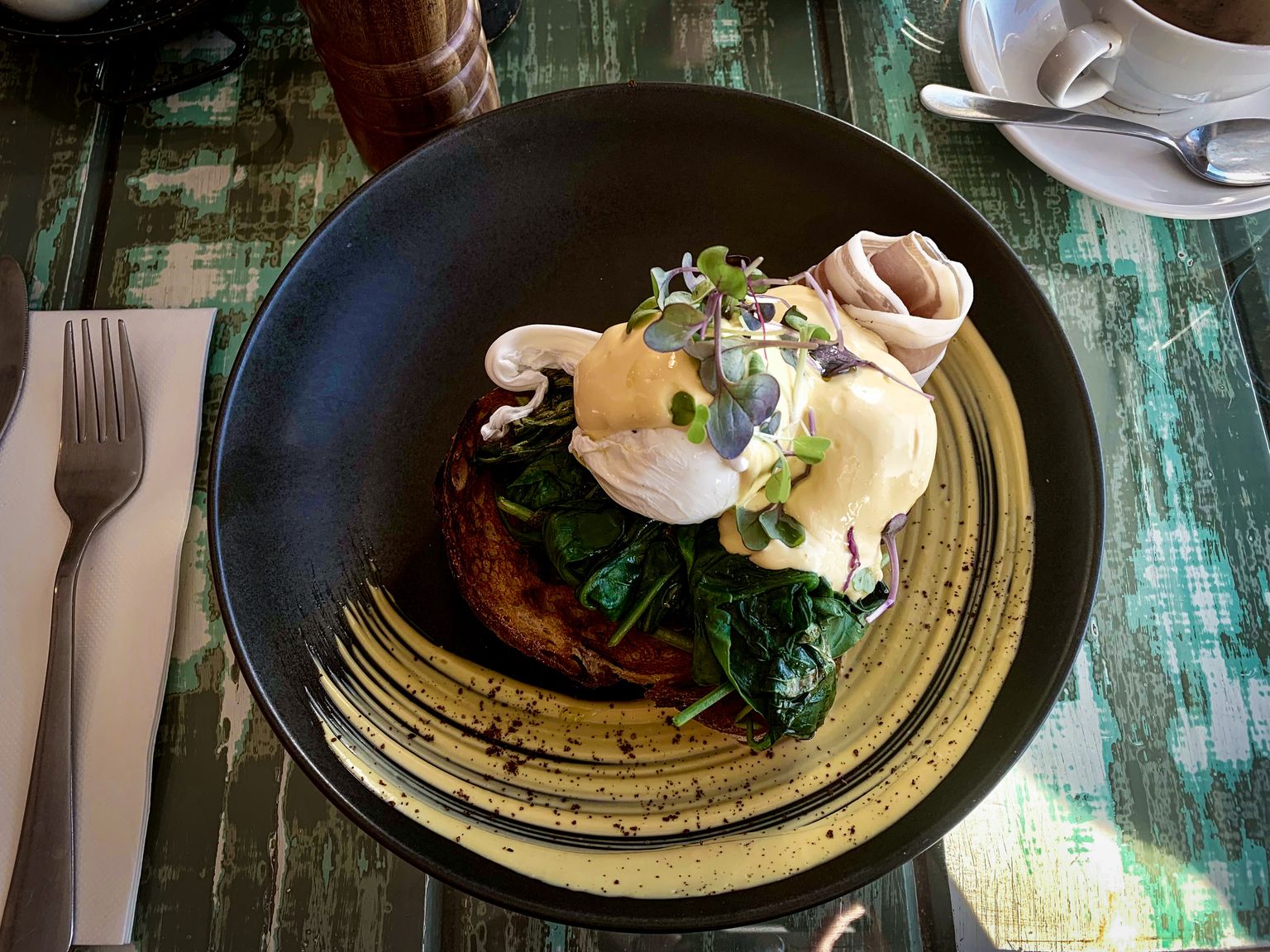 A plate of Eggs Benedict in the cafe at Foxton Beach sitting on the distressed wood table. The presentation is elegant, with the prosciutto neatly wrapped into a cylinder and the hollandaise garnished with micro greens.