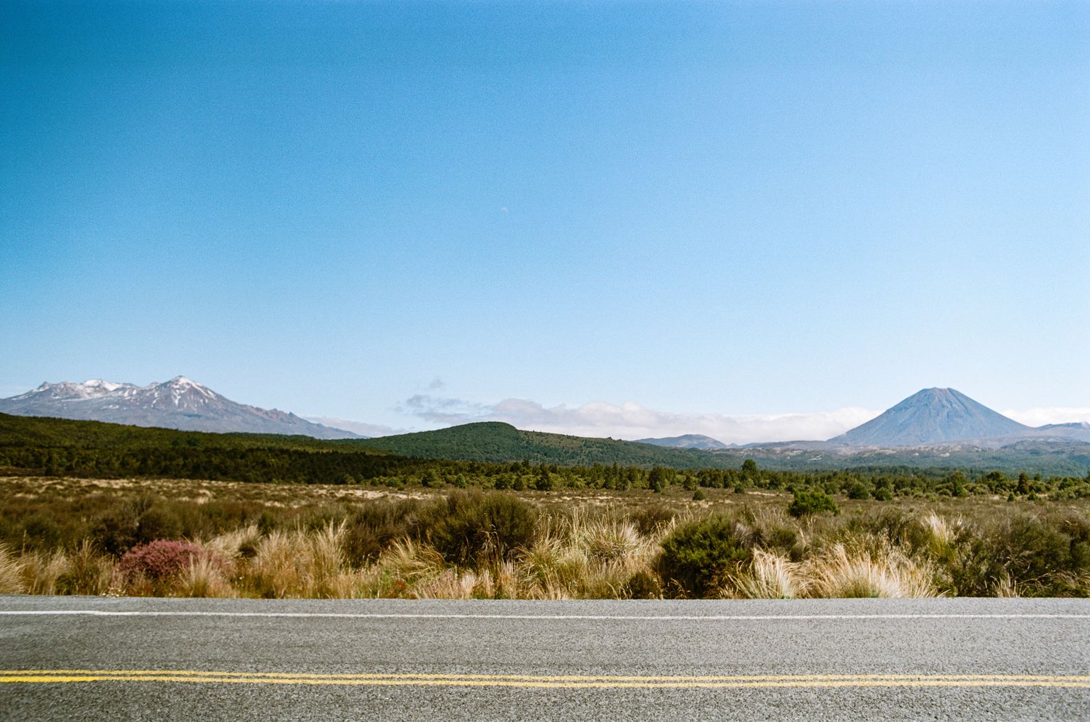 Looking across a road into a scrub desert with two mountains side-by-side in the distance, the left craggy and snow-covered, the right volcanic gray with a cinder cone.