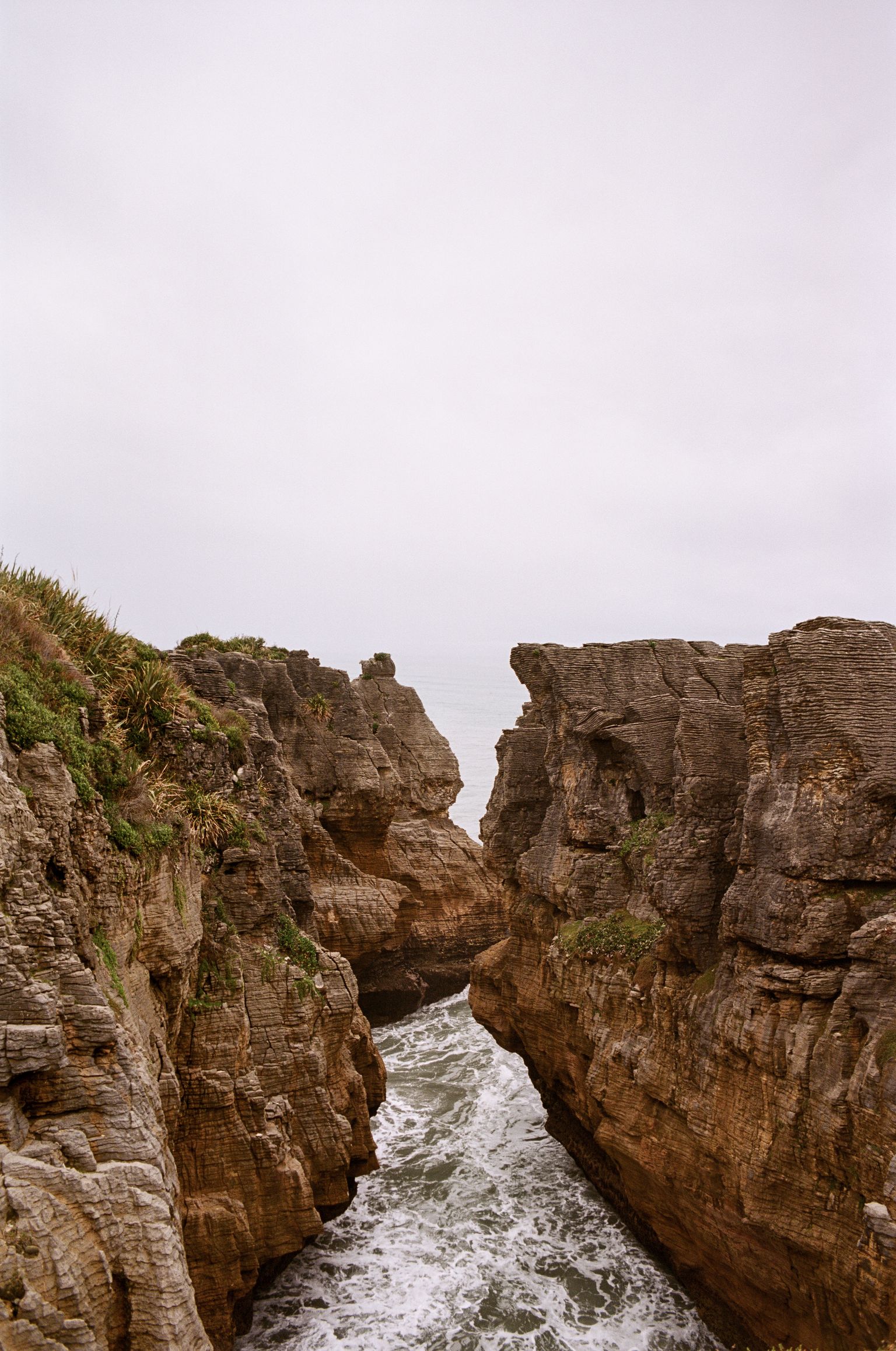 Distinctively layered rock formations jut out of the ocean and form a small canyon. The sky is hazy and overcast; the water fades white into the horizon.